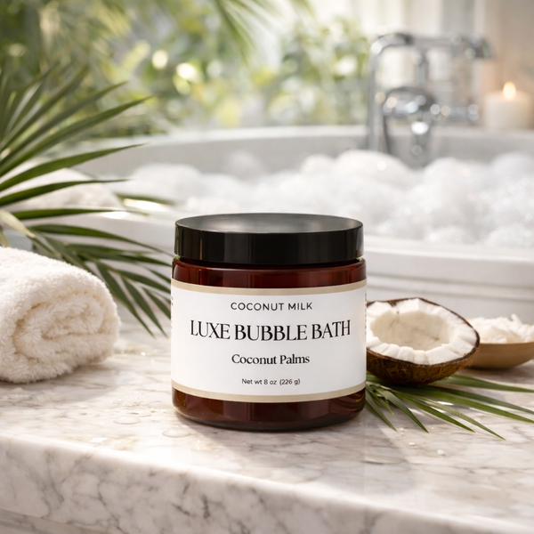 Jar of Coconut Milk Luxe Bubble Bath on a marble surface with a bathtub and palm leaves in the background.